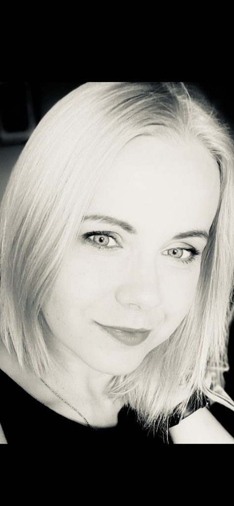Kobieta z blond włosami i łańcuszkiem na szyi uśmiecha się patrząc w stronę odbiorcy.